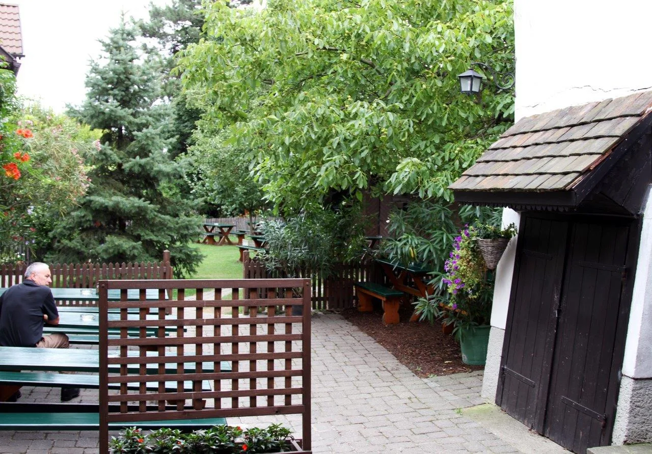 Weg in den Gastgarten, links Bänke, rechts ein Holzgebäude mit Tor